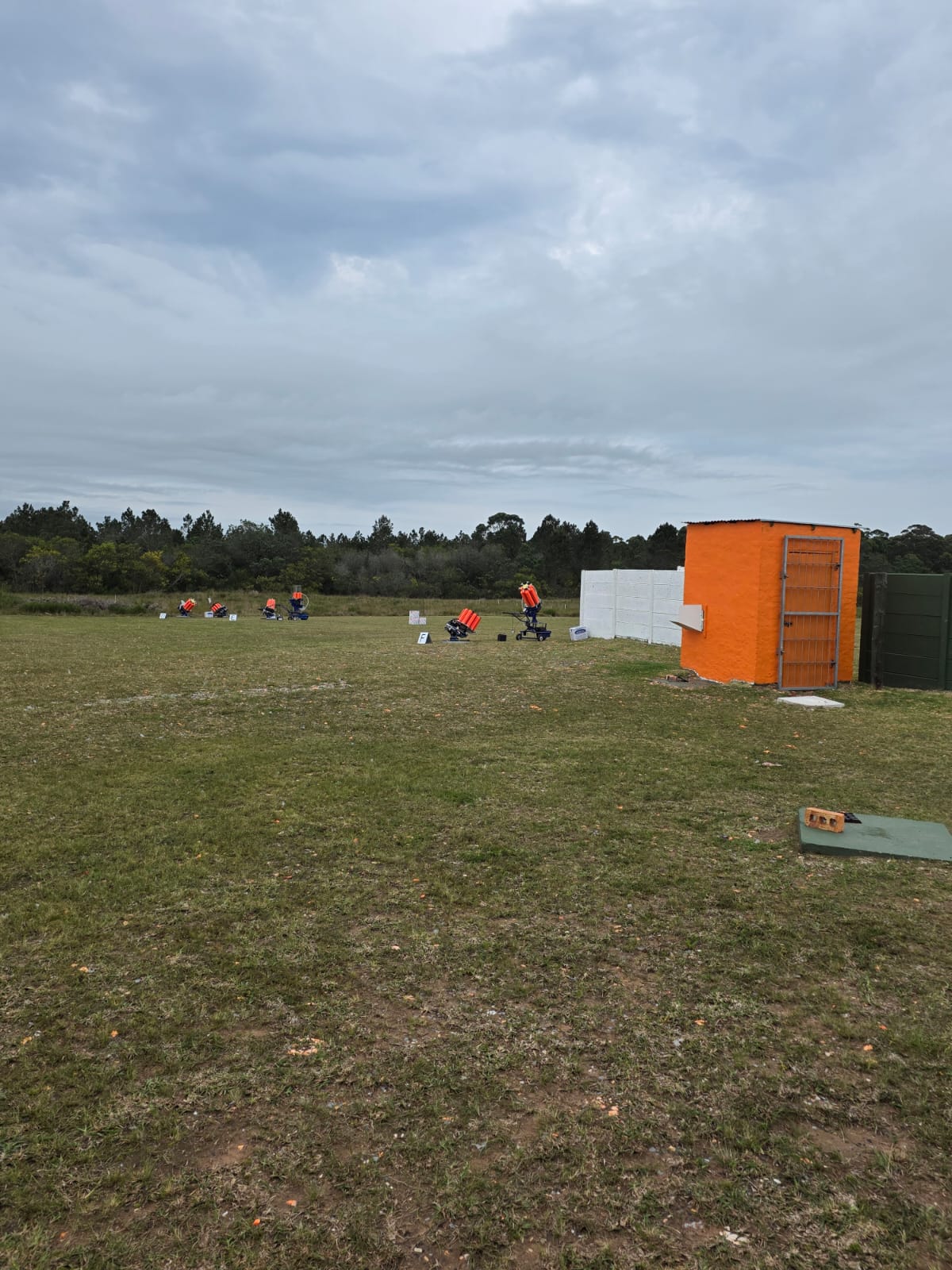 An orange trap house beside a green fence with clay target machines set out on the grass range