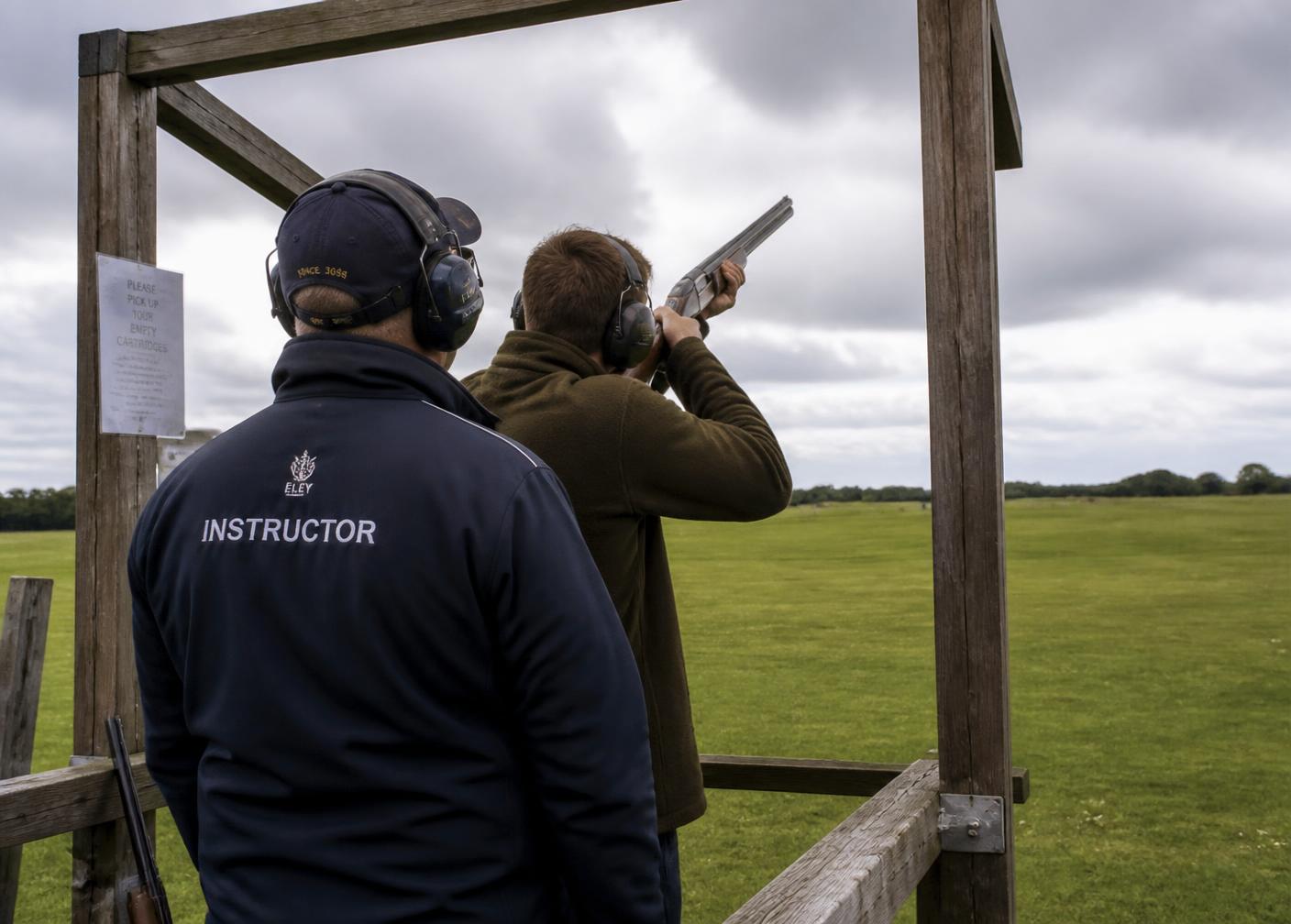 A range officer standing behind a first-time shooter at an outdoor clay shooting stand